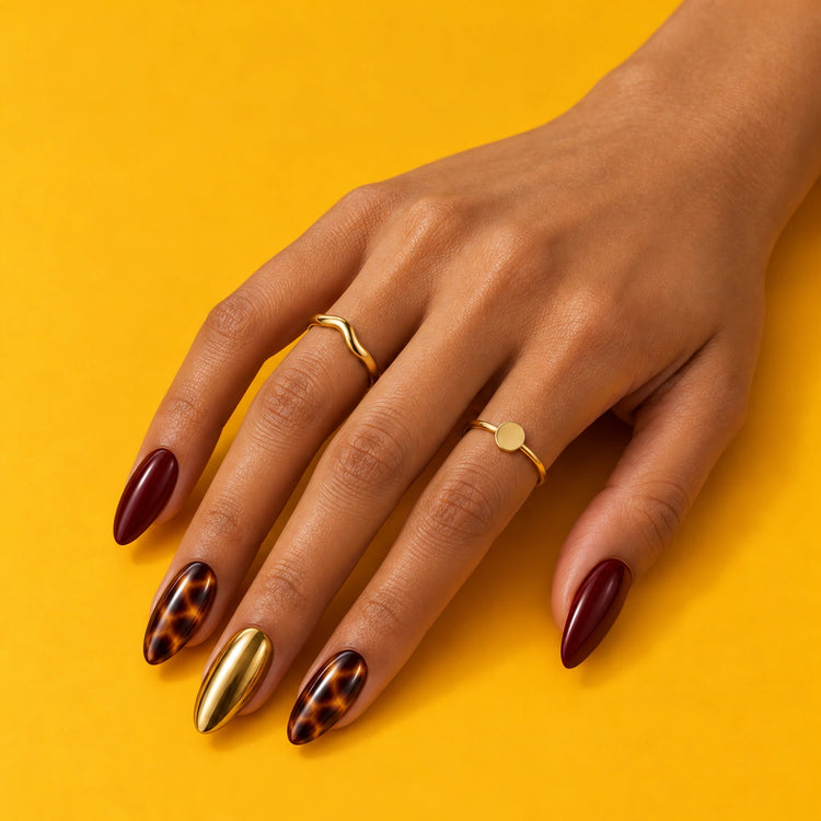 A hand on mustard yellow wearing On Me: burgundy, tortoiseshell and gold almond nails.